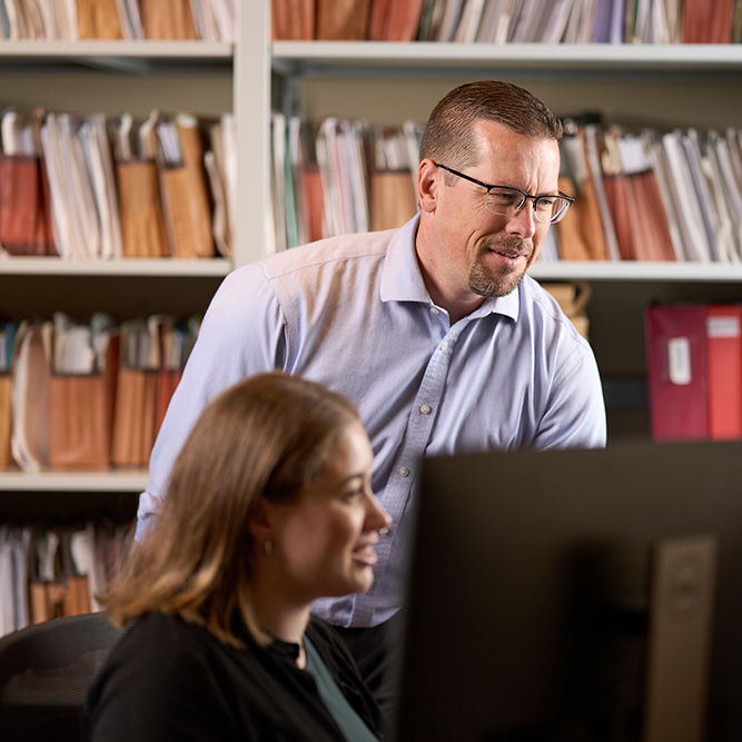 Lawyer reviewing work on a computer with a colleague, shelves of client files behind them